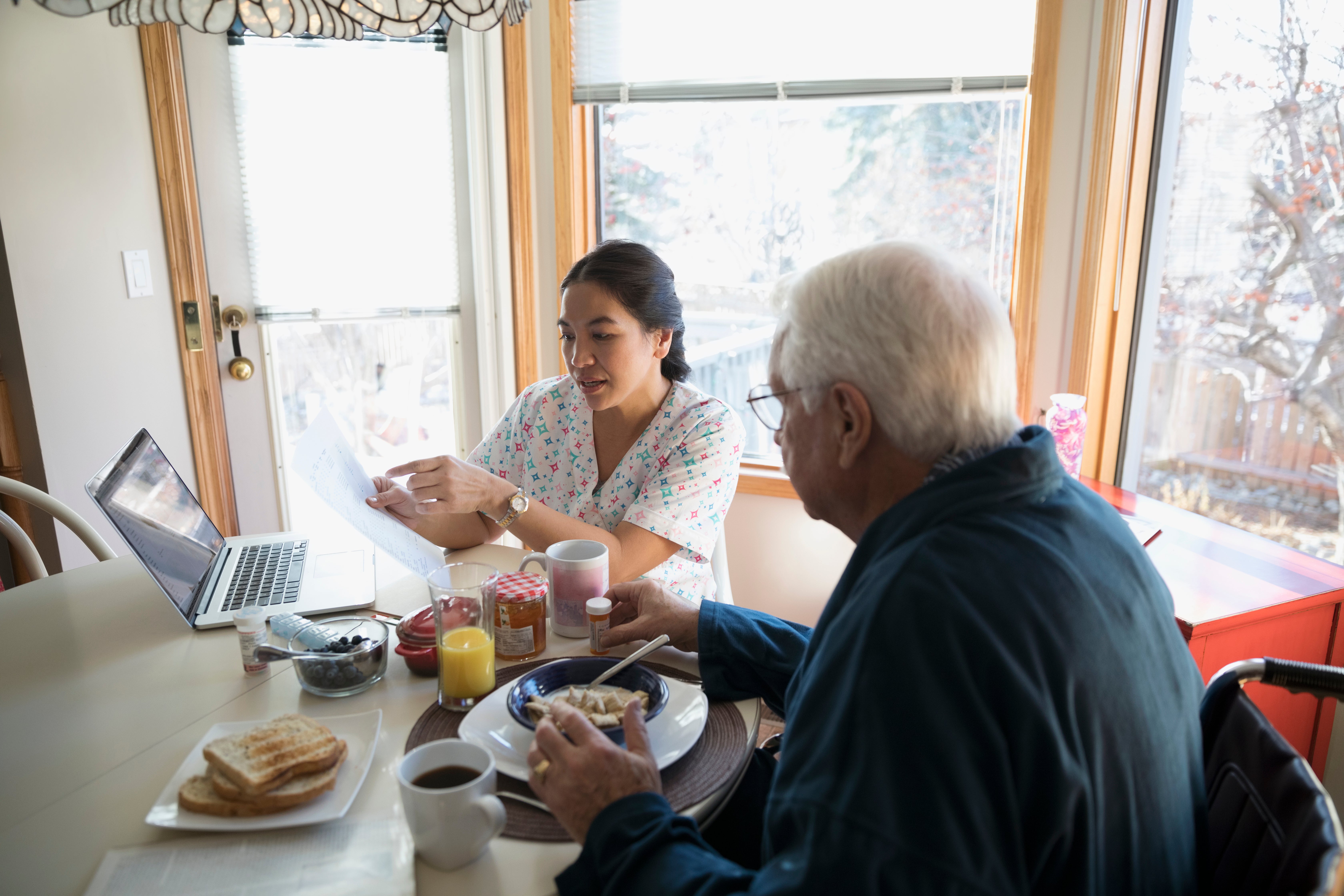 Home caregiver explaining paperwork to senior man eating breakfast at dining table