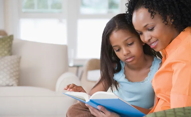 Side profile of a mid adult woman reading a book with her daughter and smiling