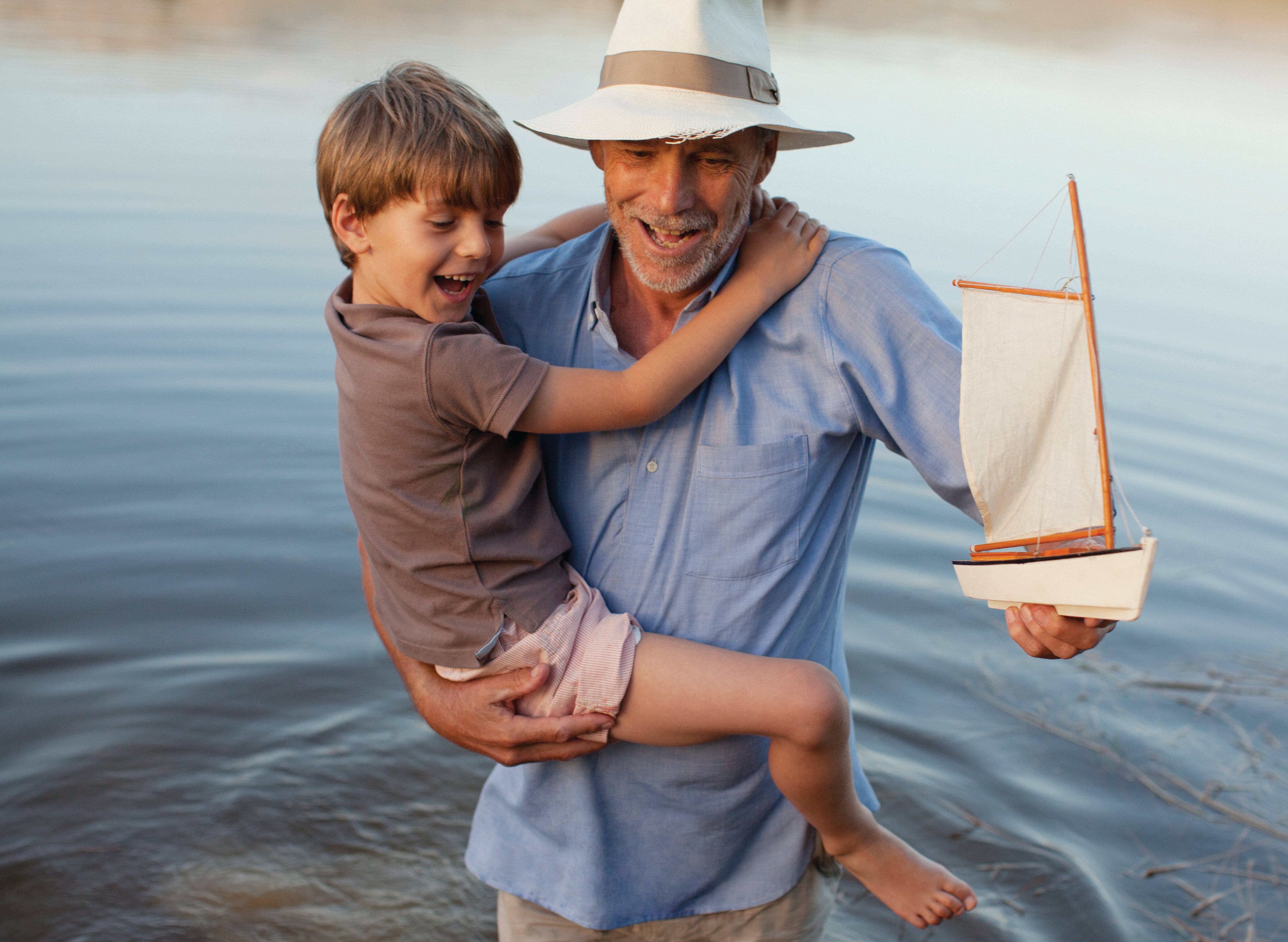 Smiling grandfather and grandson with toy sailboat wading in lake