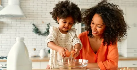 Black mother and toddler daughter in a kitchen cracking eggs into a glass bowl.