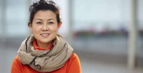 Smiling Asian woman wearing an orange sweater and beige scarf wrapped around her neck is causally standing with her arms crossed. 