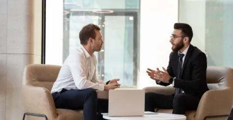 Two millennial men in suits sitting down and having an engaging conversation 


