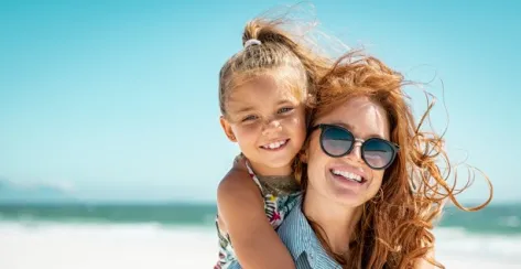 Smiling mother in sunglasses with her young daughter embracing her on a sunny day at the beach.