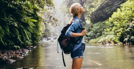 Profile view of a relaxed young female hiker with a backpack standing by a stream surrounded by trees.