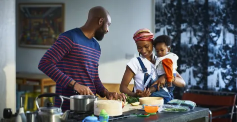 Black mother holds her baby in arms while smiling next to her husband and cooking in the kitchen.
