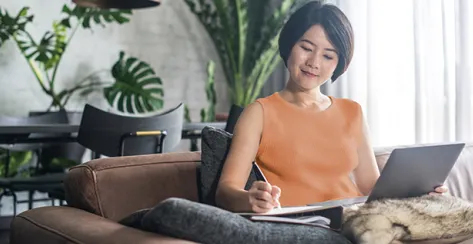 Asian woman taking notes on paper while sitting on her couch at home.