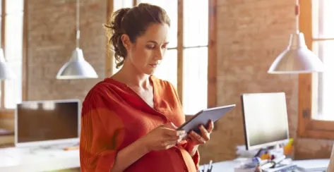 Pregnant brunette-haired woman standing in an office setting wearing an orange blouse reading a tablet