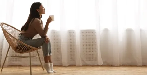 Casually dressed, young Asian woman enjoys a mug of coffee in a wicker chair near a window with a white curtain.
