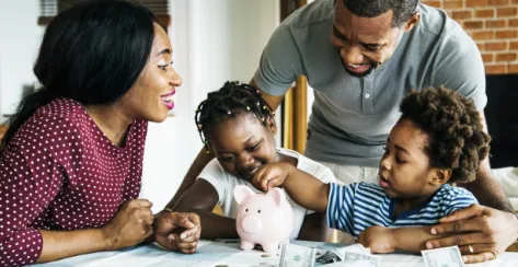 Black family laughing and sitting at the dinner table with a piggy bank and money on the counter.