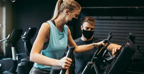 Young fit and attractive woman at body workout in modern gym together with her personal fitness instructor or coach. They keeping distance and wearing protective face masks. Coronavirus world pandemic and sport theme.