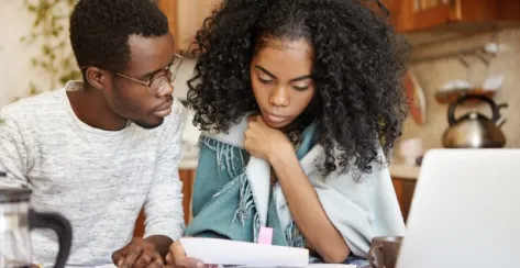 Casually dressed, young Black couple review bills together at their kitchen table with an open laptop