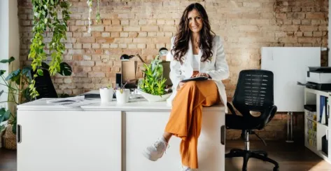 A confident woman in casual business attire with her legs crossed sits on a desk in a modern office